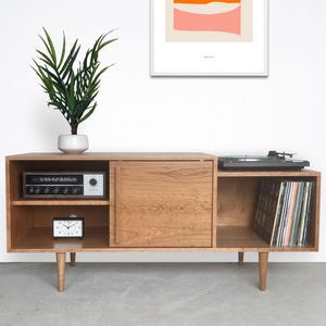 May include: A mid-century modern style wooden media console with a built-in turntable, radio, and storage for vinyl records. The console has a light wood finish, a sliding door, and tapered legs. A small clock and a potted plant are also visible.