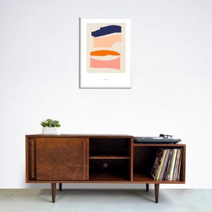 May include: A mid-century modern style wooden entertainment center with two sliding doors, two shelves, and a record player on the right side. The entertainment center is brown and has a record player on top of the right side. The record player is black and has a stack of colorful vinyl records on the shelf below it. A framed abstract art print with blue, orange, and pink shapes hangs on the wall above the entertainment center.