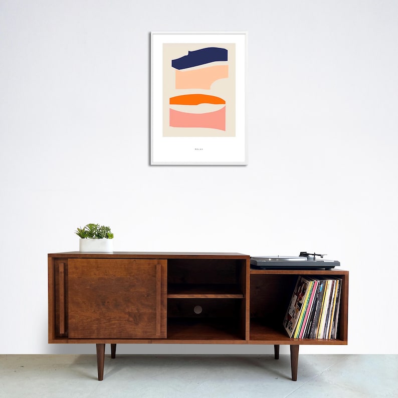 May include: A mid-century modern style wooden entertainment center with two sliding doors, two shelves, and a record player on the right side. The entertainment center is brown and has a record player on top of the right side. The record player is black and has a stack of colorful vinyl records on the shelf below it. A framed abstract art print with blue, orange, and pink shapes hangs on the wall above the entertainment center.
