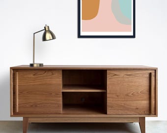Könnte beinhalten: Eine Holz-TV-Konsole mit zwei Schiebetüren und einem Regal. Die Konsole hat eine hellbraune Farbe und steht auf einem hellgrauen Boden. Eine Tischlampe aus Messing befindet sich an einer weißen Wand hinter der Konsole.
