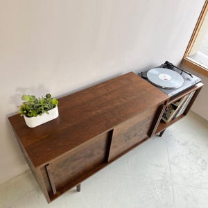 May include: A dark brown wooden record player stand with sliding doors and a shelf for vinyl records. The stand has a turntable on top and is sitting on a light gray floor.