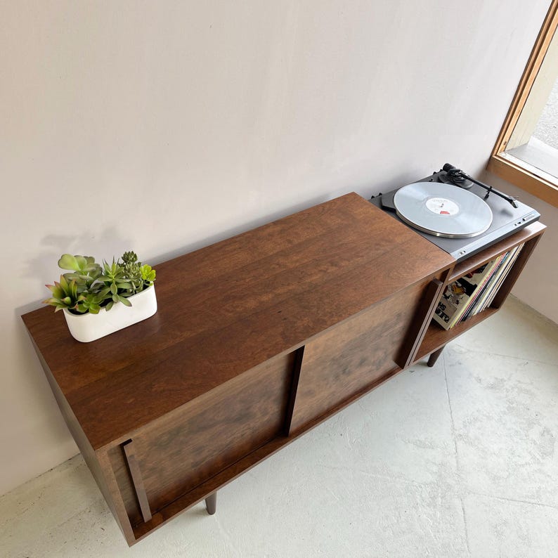 May include: A dark brown wooden record player stand with sliding doors and a shelf for vinyl records. The stand has a turntable on top and is sitting on a light gray floor.