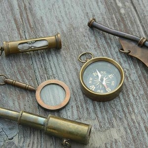 May include: An assortment of antique brass nautical instruments, including a compass, telescope, hourglass, magnifying glass, and a whistle with an anchor emblem. The items are arranged on a weathered wooden surface.