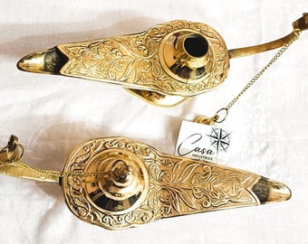 May include: Two ornate, gold-colored Aladdin lamps with intricate floral patterns. Each lamp features a spout, a handle, and a chain. The lamps are displayed on a white surface, with a tag that reads "Casa Industries".