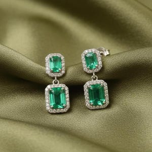 May include: A pair of emerald-cut emerald earrings with a halo of small, clear stones. The earrings are set in a silver metal and hang from a post. The background is a draped, olive-green fabric.
