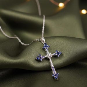 May include: A silver cross pendant necklace with blue and clear gemstones. The cross features a detailed design with a central row of clear stones and blue stones at the ends of each arm. The necklace is displayed on a green fabric.