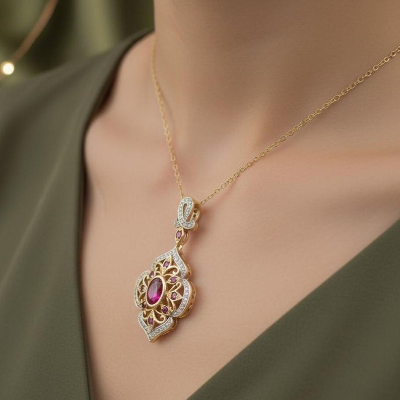 May include: A gold-colored pendant necklace with a large oval ruby center stone. The pendant has an ornate design with smaller ruby accents and clear stone embellishments. The necklace is worn against a green background.
