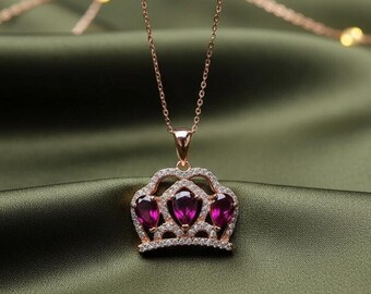 May include: A rose gold necklace with a crown pendant. The crown features three teardrop-shaped, deep pink gemstones surrounded by small, clear crystals. The necklace chain is delicate, and the pendant is centered on a green satin background.