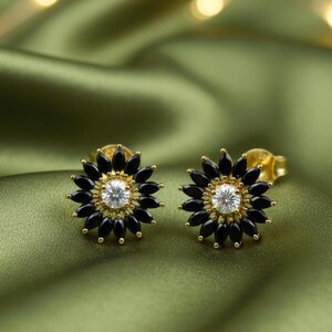 May include: A pair of gold-coloured stud earrings. Each earring has a central, round, clear gemstone surrounded by black, petal-shaped stones. The earrings are set against a green satin background.