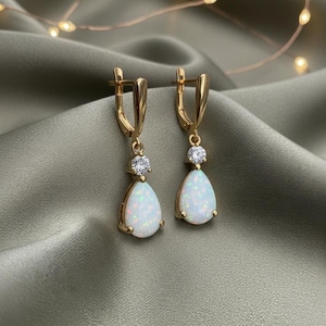 May include: A pair of gold-colored drop earrings. Each earring features a pear-shaped, white opal stone, accented with a small, round, clear gemstone above. The earrings have a lever-back closure. The background is a draped, olive-green fabric.