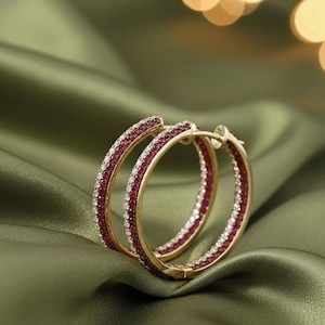 Puede incluir: Un par de pendientes de aro dorados con piedras preciosas rojas y blancas. Los pendientes están colocados sobre un fondo de satén verde, con luces suaves y borrosas en el fondo.