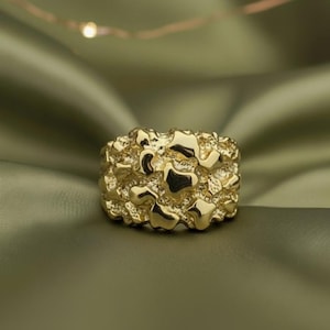 Op de afbeelding: Een goudkleurige ring met een getextureerd, nugget-achtig ontwerp. De ring heeft een onregelmatig oppervlak met verschillende vormen en maten, wat een unieke uitstraling creëert. De ring is een opvallend sieraad.