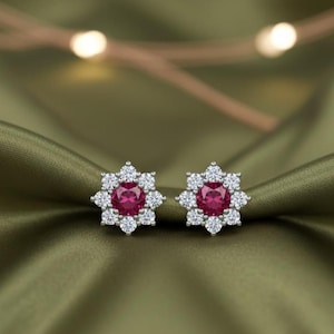 Może przedstawiać: Para kolczyków sztyftów z centralnym, okrągłym, głęboko różowym kamieniem szlachetnym, otoczonym mniejszymi, przezroczystymi, okrągłymi kamieniami szlachetnymi. Kolczyki są osadzone w srebrnym metalu i prezentowane na zielonej, satynowej tkaninie.