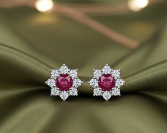 May include: A pair of stud earrings featuring a central, round, deep pink gemstone surrounded by a halo of smaller, clear, round gemstones. The earrings are set on a silver-colored metal and are displayed on a green, satin-like fabric.