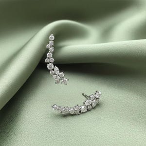 May include: A pair of silver-toned ear climbers, each adorned with a cascade of sparkling, clear gemstones. The earrings curve upwards, creating a delicate, elegant design. The background is a soft, muted green fabric.