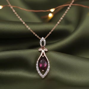 Può includere: Una collana in oro rosa con un pendente con una pietra preziosa rosso scuro a taglio marquise. Il pendente è delineato da piccole pietre bianche scintillanti. La collana è appesa a una delicata catena, su uno sfondo verde.