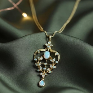 May include: A gold-toned pendant necklace with an ornate, filigree design. The pendant features an oval opal stone at the center, surrounded by smaller opals and leaf-like details. The necklace hangs on a delicate chain, set against a dark green background.