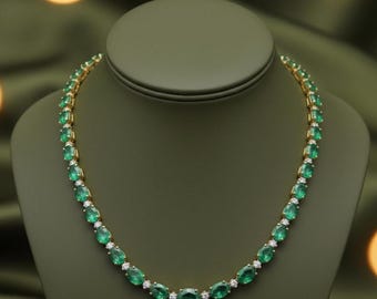 Op de afbeelding: Een gouden ketting met ovale smaragden en kleine, ronde diamanten. De ketting wordt getoond op een groene sieraden buste tegen een groene achtergrond. De smaragden zijn heldergroen.