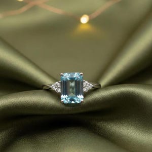 May include: A silver ring with a large, rectangular, light blue gemstone at its centre. Small, round, clear gemstones are set either side of the main stone. The ring is displayed on a green satin background, giving a luxurious appearance.