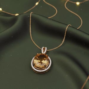 May include: A gold necklace featuring a large, oval, amber-coloured gemstone pendant. The pendant is surrounded by a halo of small, sparkling white stones. The necklace chain is delicate and gold, set against a dark green backdrop.