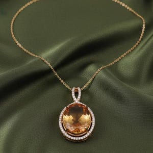 May include: A gold necklace featuring a large, oval amber gemstone pendant. The pendant is encircled by a halo of small, clear gemstones. The necklace is displayed on a green satin fabric.