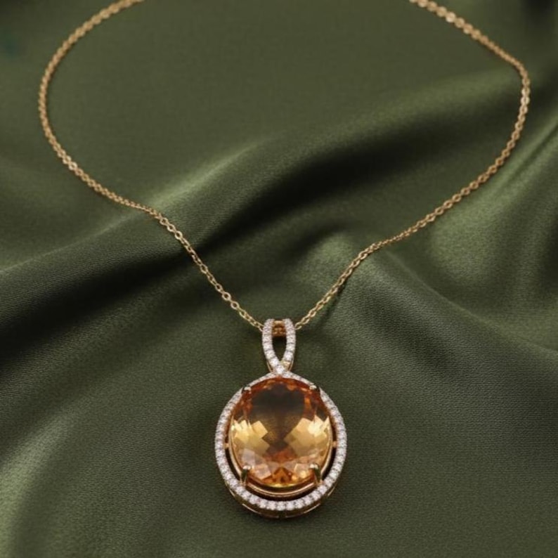 May include: A gold necklace featuring a large, oval amber gemstone pendant. The pendant is encircled by a halo of small, clear gemstones. The necklace is displayed on a green satin fabric.