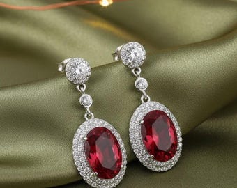 May include: Dangling silver earrings with oval, deep red gemstones surrounded by a halo of small, clear stones. The earrings have a post and three smaller stones connecting to the main gemstone. The background is a silky, olive green fabric.