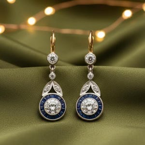 May include: A pair of drop earrings with gold-tone hooks and silver-tone settings. Each earring has a large, round, clear stone encircled by blue stones. Further clear stones and decorative elements complete the design, creating an elegant look.