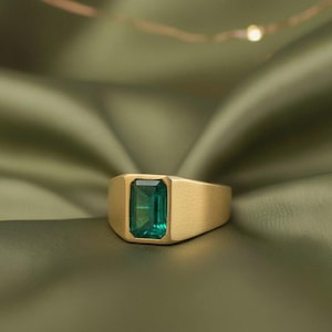 May include: A gold signet ring featuring a rectangular emerald-cut green gemstone. The ring has a brushed gold finish and a classic design. The gemstone is a vibrant green colour.