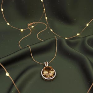 May include: A gold pendant necklace featuring a large, oval, amber gemstone, surrounded by a halo of smaller, clear stones. The necklace hangs on a fine chain against a dark green satin backdrop. Decorative fairy lights are in the background.