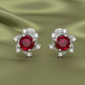 May include: A pair of stud earrings with a central, round, deep red gemstone, encircled by smaller, clear gemstones in a floral pattern. The earrings are set in silver-coloured metal. The background is a shade of green.