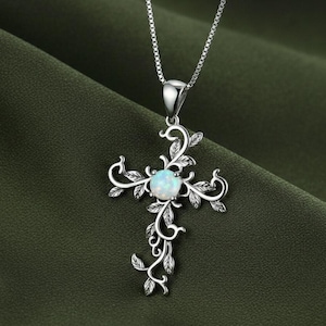 May include: A silver-colored pendant necklace featuring a cross design. The cross is adorned with a central opal-like stone and intricate leaf and vine detailing. The necklace hangs on a delicate chain, set against a dark green background.
