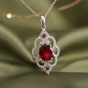 May include: A silver pendant necklace featuring a large, oval-cut ruby gemstone. The ruby is surrounded by a decorative openwork design with small, clear gemstones. Two smaller rubies accent the design. The necklace hangs on a delicate silver chain.