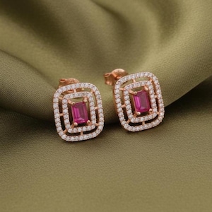 May include: A pair of rose gold stud earrings. Each earring features a rectangular, magenta-colored gemstone at the center, surrounded by a square frame of small, clear crystals. The earrings are set against a green background.