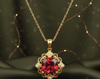 May include: A gold-colored necklace with a square, red gemstone pendant. The pendant is surrounded by a decorative gold frame with small, clear gemstones. The necklace chain is also gold and hangs against a dark green background.