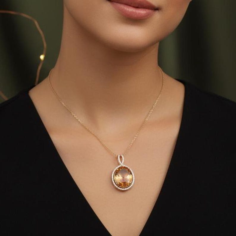 May include: A gold necklace with a large, round pendant. The pendant features a large, amber-coloured gemstone surrounded by a halo of smaller, clear stones. The necklace is worn against a black top.