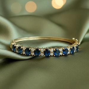 Puede incluir: Una pulsera dorada con piedras preciosas ovales de color azul oscuro y piedras preciosas redondas y claras más pequeñas. La pulsera está sobre un fondo de satén verde, con luces borrosas.