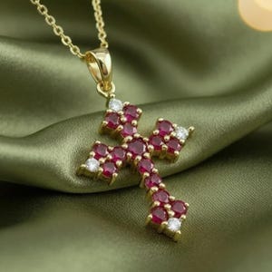 May include: A gold cross pendant necklace with a chain. The cross is set with numerous round, deep red gemstones and small, clear accent stones. The necklace is displayed on a green, textured fabric.