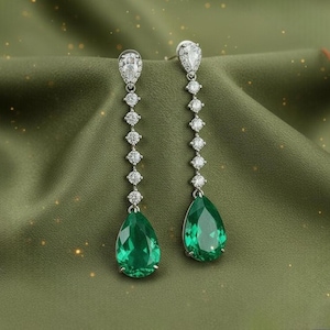 May include: A pair of elegant drop earrings featuring a teardrop-shaped emerald green gemstone. The earrings have a silver-colored setting with a series of small, round, clear gemstones. The earrings are displayed on a green fabric background.