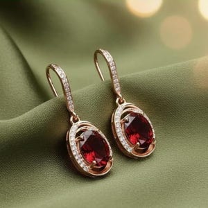 May include: A pair of rose gold drop earrings with oval, deep red gemstones. Each earring features a halo of small, clear crystals around the gemstone and along the top hook. The earrings are displayed on a green fabric.