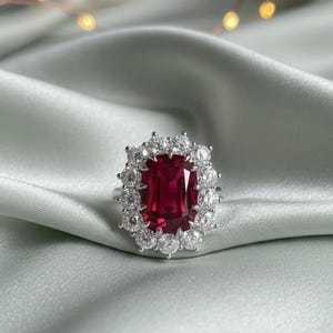 May include: A silver ring featuring a large, rectangular, deep red gemstone surrounded by a halo of smaller, clear gemstones. The ring is set against a soft, light green fabric, creating a luxurious feel.