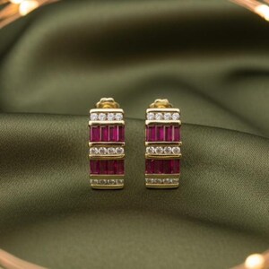 May include: A pair of gold-toned earrings with a rectangular design. Each earring features rows of small, rectangular, ruby-colored stones and clear, round accent stones. The earrings are set against a green background.