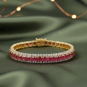 Puede incluir: Una pulsera dorada con una fila de piedras rojas cuadradas y una fila de pequeñas piedras blancas. La pulsera está sobre un fondo verde.