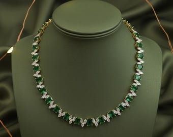 Op de afbeelding: Een goudkleurige ketting met smaragdgroene edelstenen en kleine witte accentstenen. De ketting wordt getoond op een groene sieraden buste tegen een donkergroene achtergrond. De edelstenen zijn in een herhalend patroon gerangschikt.