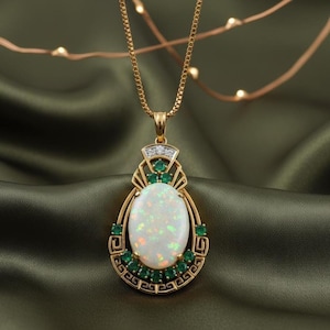 Könnte beinhalten: Goldfarbene Halskette mit Anhänger, besetzt mit einem ovalen Opal-Edelstein, kleinen Smaragden und Diamanten. Der Anhänger hat ein kunstvolles, vom Art Déco inspiriertes Design. Die Halskette wird auf grünem Satin präsentiert.