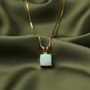 Könnte beinhalten: Eine goldene Halskette mit einem quadratischen Opalanhänger. Der Opal hat eine weiße Basis mit rosa und grünen Blitzen. Die Halskette wird auf grünem Satin präsentiert.