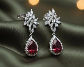 May include: A pair of silver earrings with a floral design and teardrop-shaped red gemstones. The earrings have a halo of small, clear stones around the red gems. The background is a dark green fabric.