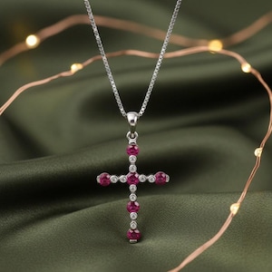 May include: A silver-toned pendant necklace featuring a cross design. The cross is adorned with round, deep red gemstones and small, clear accent stones. The necklace hangs on a delicate chain against a green background.