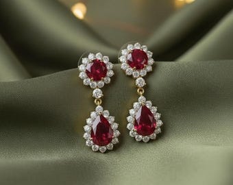 May include: A pair of gold-toned drop earrings with a floral design at the top and a teardrop shape at the bottom. Each earring features a large, deep red gemstone surrounded by small, clear crystals. The earrings are displayed on a green fabric.