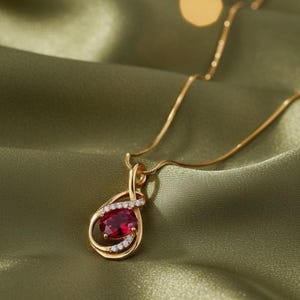 Puede incluir: Un collar con colgante dorado con una piedra preciosa roja ovalada y pequeñas piedras blancas. El colgante cuelga de una delicada cadena dorada, sobre un fondo de satén verde. La joya es elegante y llamativa.